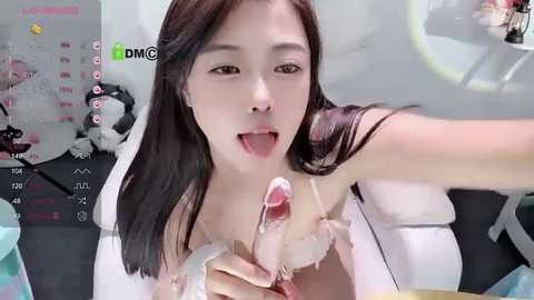 Media: Video of an East Asian woman with long dark hair, licking a man's erect penis, wearing a white lace bra. Background features a gaming screen with digital text and icons.