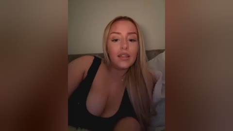 Media: Video of a blonde woman with fair skin and medium breasts, wearing a low-cut black tank top, reclining on a bed with white pillows.