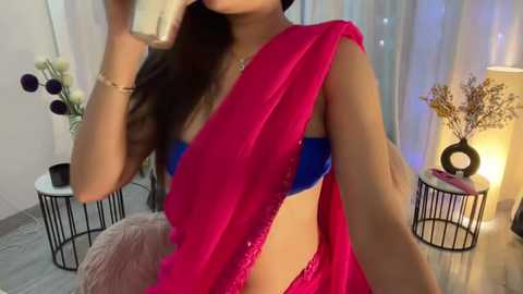 Media: Video of a South Asian woman with long dark hair, wearing a vibrant pink sari and blue lingerie, holding a glass. Background includes a white wall, floral arrangements, and a lamp.