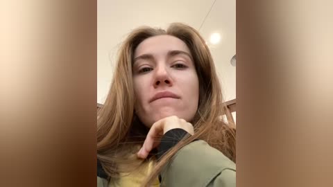 Media: A close-up video of a fair-skinned woman with long brown hair, wearing a green jacket, looking introspective, taken indoors with a blurred background.