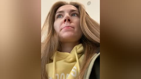 Media: Video of a young woman with long, straight brown hair, wearing a yellow hoodie, gazing upward with a thoughtful expression. The background is blurred and neutral-toned.