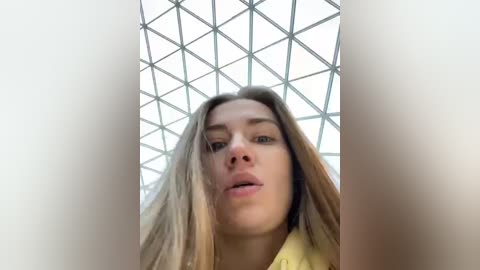 Media: Video of a young woman with long, straight blonde hair, fair skin, and light makeup, looking slightly upward with parted lips. She's wearing a yellow top. Background shows a geometric, grid-patterned ceiling.