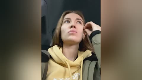 Media: Video of a young woman with light skin and straight, shoulder-length brown hair, wearing a yellow hoodie and green jacket. She's gazing up thoughtfully, with a hand touching her chin. The background is blurred.