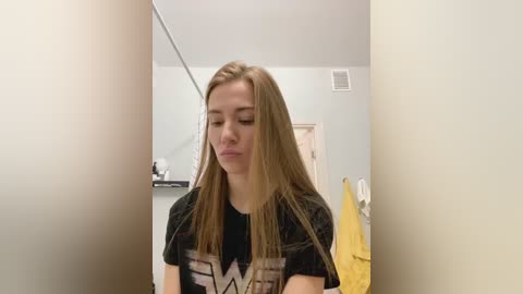 Media: Video of a young Caucasian woman with long, straight blonde hair, wearing a black graphic T-shirt, standing indoors with a light blue wall and a yellow towel in the background.