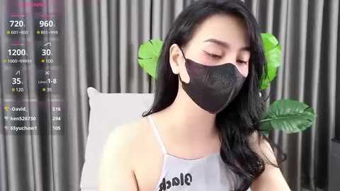 Media: Video of an Asian woman with long black hair, wearing a black face mask, grey tank top, and green plant behind her, displaying social media stats on the left.