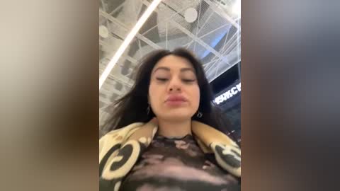 Media: Video of a woman with long black hair, fair skin, and full lips, wearing a floral blouse, against a modern ceiling with circular patterns and a partially visible \"Sephora\" sign.