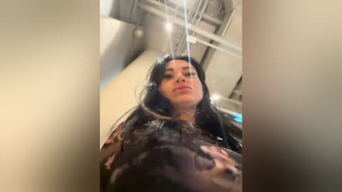 Media: Video of a woman with long black hair, wearing a black leather jacket with a distressed design, standing in a dimly lit, industrial setting with exposed pipes and a metal ceiling.