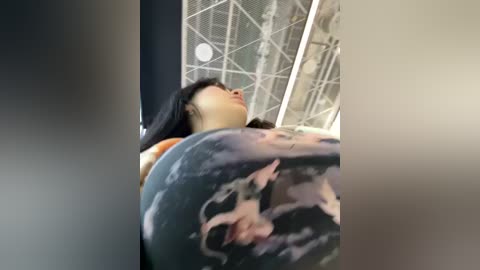 Media: Video of a woman with long black hair wearing a graphic print shirt, seen from a low angle, with a metal structure and light source in the background.