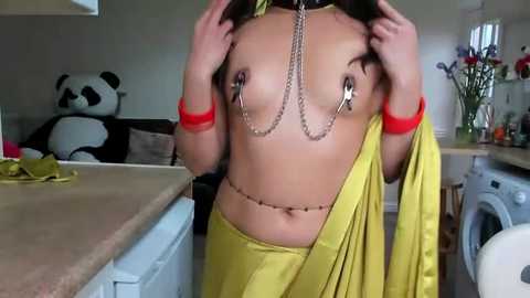 Media: Video of a topless woman with medium breasts, wearing a yellow sari and red bangles, holding her hair up, in a modern kitchen with a panda toy, flowers, and a washer.