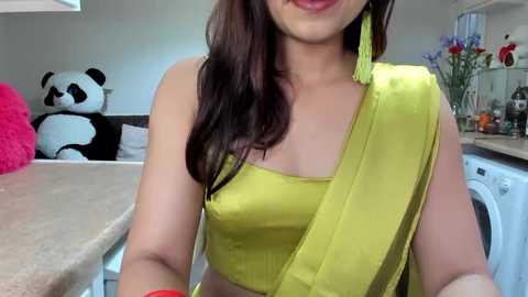 Media: Video of a smiling South Asian woman in a mustard yellow saree, large pink flower in background, white washing machine, kitchen sink, and a stuffed panda toy.