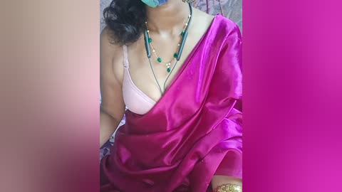 Media: Video of a woman with medium skin tone, wearing a pink bra, draped in a vibrant magenta sari, adorned with green beads, and a teal face mask.