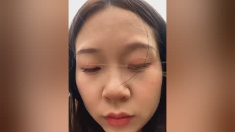 Media: Video of an Asian woman with fair skin, dark hair, and closed eyes, with a noticeable scar on her forehead. The background is blurred, focusing attention on her facial features.