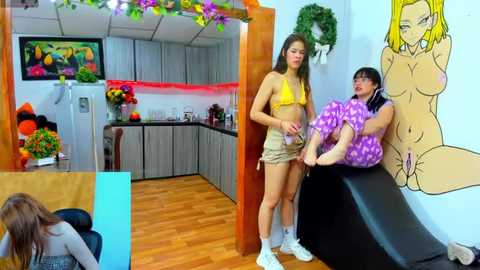 Media: Video of two young women, one in a yellow bikini, the other in purple polka-dot pajamas, in a vibrant, cartoon-themed kitchen with a large, nude anime girl mural.