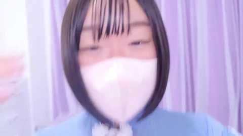 Media: Video of a person with short, straight black hair wearing a white surgical mask, standing against a blurred lavender background. The image is slightly out of focus.