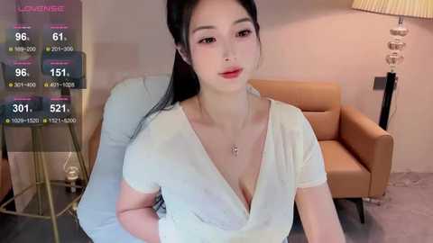 Media: A video of an East Asian woman with long black hair, fair skin, and red lipstick, wearing a low-cut white top, sitting in a beige armchair. Background includes a lamp, a wooden floor, and a green plant.