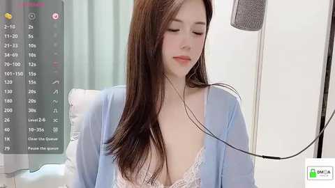 Media: Video of a young Asian woman with long brown hair, wearing a light blue robe over a lacy white bra, seated in a medical setting with monitors and IV equipment.