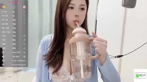 Media: Video of an Asian woman with long brown hair, wearing a light blue cardigan and a lace bra, drinking from a baby bottle, in a modern, clinical setting.