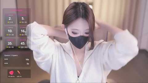 Media: A video of an Asian woman with light skin, wearing a white long-sleeve top and black face mask, adjusting her headphones while indoors with beige curtains.