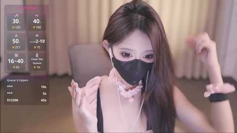 Media: A video of an East Asian woman with long, straight brown hair, wearing a black face mask, a pink ruffled blouse, and white headphones, sitting indoors with beige curtains in the background.