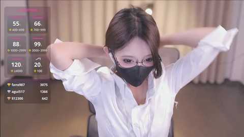 Media: Video of a young East Asian woman with straight black hair, wearing a white blouse, black face mask, and glasses, stretching her arms behind her head in a bright, modern office setting.