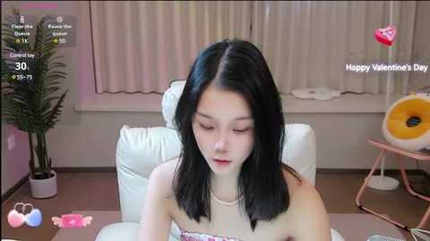 Media: Video of an Asian woman with straight black hair and fair skin, wearing a strapless floral dress, sitting on a white leather chair in a cozy room. Background includes a potted plant, pink heart-shaped object, and Valentine's Day-themed text.