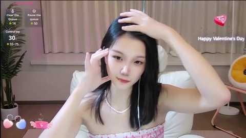 Media: Video of an East Asian woman with straight black hair, fair skin, and wearing a strapless floral top. She's seated indoors, smiling, with a pink phone and a pink heart-shaped cushion in the background.