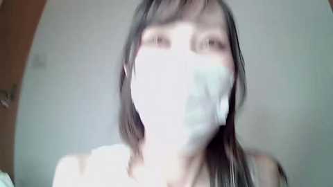 Media: A blurred video of a young Asian woman with long black hair, wearing a white face mask, standing indoors with a plain white background.