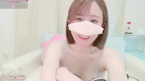 Media: Video of a young, fair-skinned woman with straight, shoulder-length brown hair, wearing a pink surgical mask and a light pink bra. She's smiling, lying on a fluffy white bed, with a soft pink blanket and a blue pillow in the background.