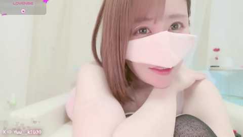 Media: A video of an Asian woman with straight, shoulder-length brown hair, wearing a pink mask, lying on a bed in a white room.