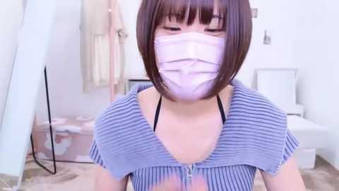 Media: Video of an Asian woman with short, straight brown hair and a surgical mask covering her nose and mouth, wearing a striped, cropped top. Background features a white room with a chair, clothes rack, and wooden floor.