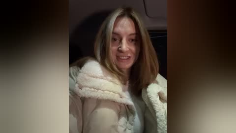 Media: Video of a smiling Caucasian woman with shoulder-length, light brown hair, wearing a white, fluffy bathrobe, with a blurred, dark background.