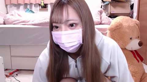 Media: Video of an Asian woman with long brown hair, wearing a light blue hoodie and a pink mask, kneeling in a cluttered bedroom with a teddy bear and bed in the background.