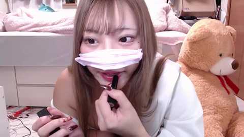 Media: A video of a young East Asian woman with light skin and straight, shoulder-length blonde hair, wearing a white mask, applying lipstick. She sits on a bed in a cluttered bedroom with a stuffed teddy bear.