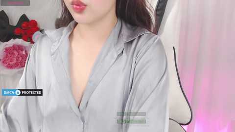 Media: Video of a woman with fair skin, wearing a loose, grey button-up shirt, lips painted in a bold red. Background features a floral arrangement and a white chair, with a watermark indicating \"DANCA PROTECTED.\