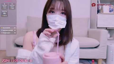 Media: Video of an Asian woman with straight brown hair and a surgical mask, wearing a white robe, seated on a couch, holding a pink cup, in a modern, minimalist living room.