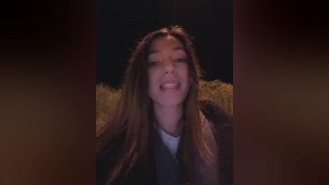 Media: A video of a young woman with long brown hair, smiling, in a dark field with golden hay in the background.