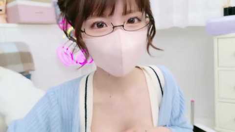 Media: Video of an Asian woman with fair skin, wearing glasses, a light blue cardigan, and a pink face mask, sitting in a brightly lit room with white walls and a pastel-colored bed.