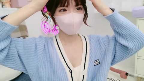 Media: Video of an Asian woman with fair skin, wearing a light blue knitted cardigan, pink surgical mask, and pink hair accessories, adjusting her hair in a white room.