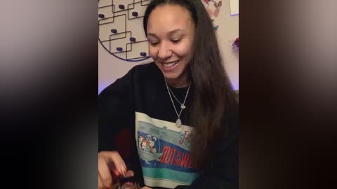 Media: Video of a smiling young woman with long dark hair, wearing a graphic tee and silver necklace, seated in a cozy room with a blackboard wall.