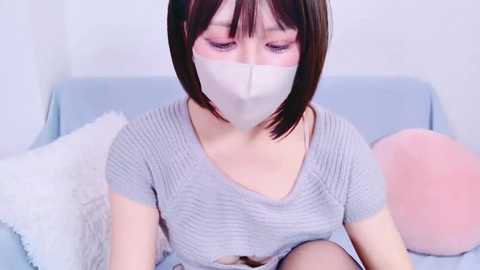 Media: Video of a young Asian woman with straight, shoulder-length black hair, wearing a light gray ribbed knit top and white face mask, sitting on a light blue couch with fluffy pillows, indoors with white walls.