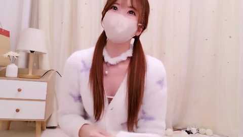 Media: Video of an Asian woman with long brown pigtails, wearing a white mask, robe, and choker, seated indoors near a white nightstand with a lamp.