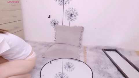 Media: A video of a white woman with blonde hair in a white shirt, kneeling on a light gray floor, installing a round mirror with a decorative flower pattern on a white wall.