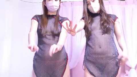 Media: Video of two East Asian women in see-through black mesh dresses, making a peace sign, in a pastel-colored room with white curtains.