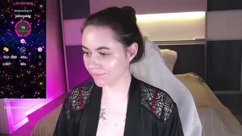 Media: Video of a fair-skinned woman with dark hair in an updo, wearing a black lace robe, a white veil, and a tattoo on her collarbone, in a modern bedroom with a digital clock and bed.