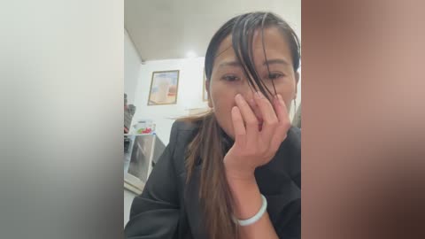 Media: A video shows an Asian woman with long, wet hair, covering her face with one hand, wearing a black robe. The background features a white wall with framed art and a shelf with items.