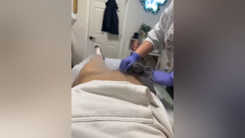 Media: A video captures a spa treatment in progress. A person lies on a massage table, draped in a white towel, while a therapist in purple gloves works on their lower back. The room is bright with a white door, a blue jacket hanging, and a plant in the background.