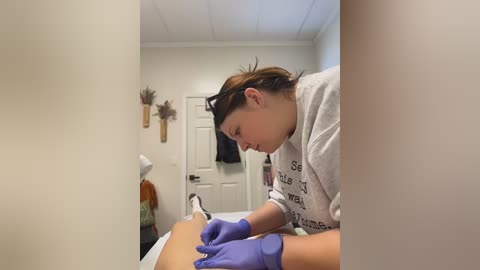 Media: Video of a woman with brown hair, wearing a gray T-shirt, purple gloves, and a headband, applying a tattoo on a client's leg in a clean, white-walled room with a door, shelves, and a white ceiling.