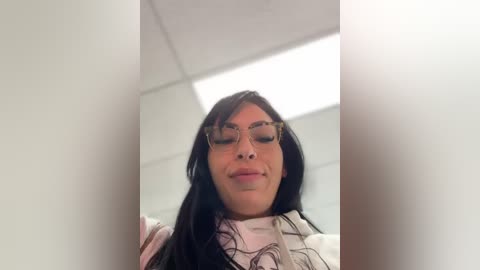 Media: Video of a smiling woman with long black hair, wearing round glasses, a white top, and a gray sweater, standing in a brightly lit, white-tiled bathroom with fluorescent lighting.