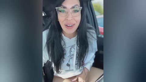 Media: A video of a woman with long black hair, wearing glasses and a white t-shirt with a graphic design, smiling inside a car.