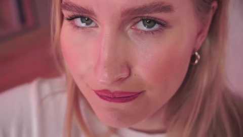 Media: Video of a fair-skinned, blonde woman with green eyes, wearing subtle makeup, and a white top. She has a neutral expression, with a hint of a smile. The background is blurred, featuring warm tones.